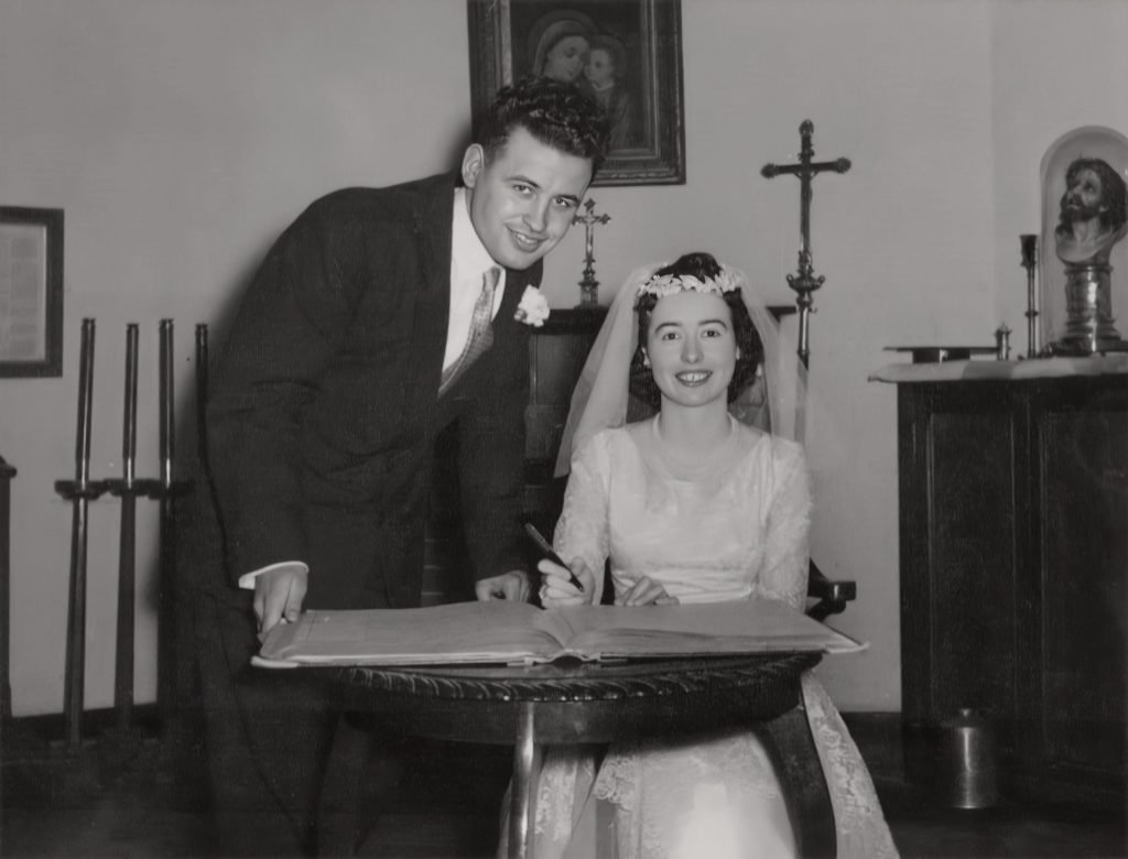 Black-and-white photo of a bride in a veil signing the register beside her groom in a suit, smiling at the camera in a church sacristy with religious statues and a crucifix behind them.