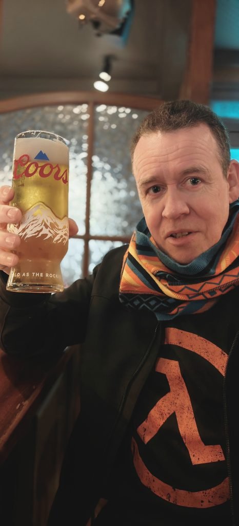A man in a patterned scarf raises a pint of Coors toward the camera, seated at a wooden bar table with a frosted window lit up behind him.