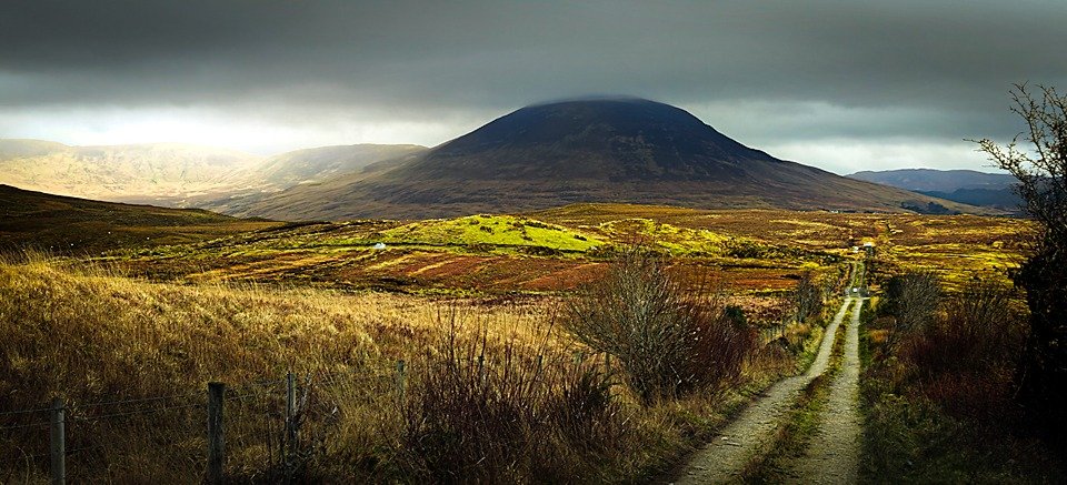 A dirt track leads through autumn-coloured bogland toward a dark, rounded mountain, its summit wrapped in cloud under a moody sky.