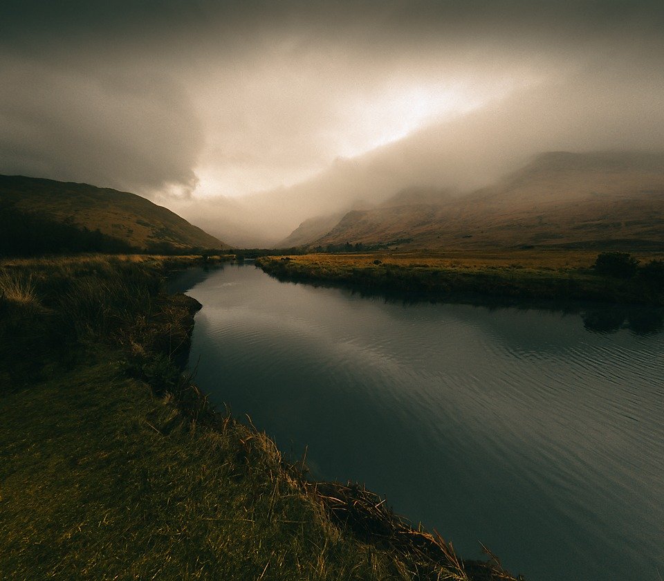 A still river winds between grassy banks toward mist-shrouded hills, with low storm light breaking through heavy cloud over the valley.