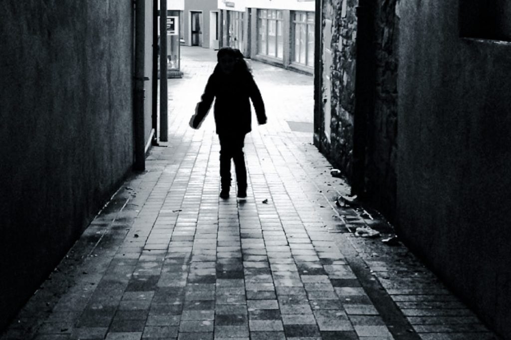 Black-and-white photo of a man's silhouette walking down a narrow laneway toward a bright street beyond, flanked by dark stone walls.