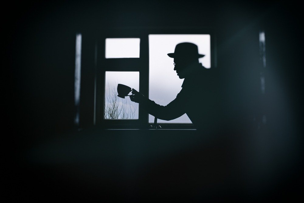Silhouette of a person in a hat holding a cup, framed by a window at dusk.