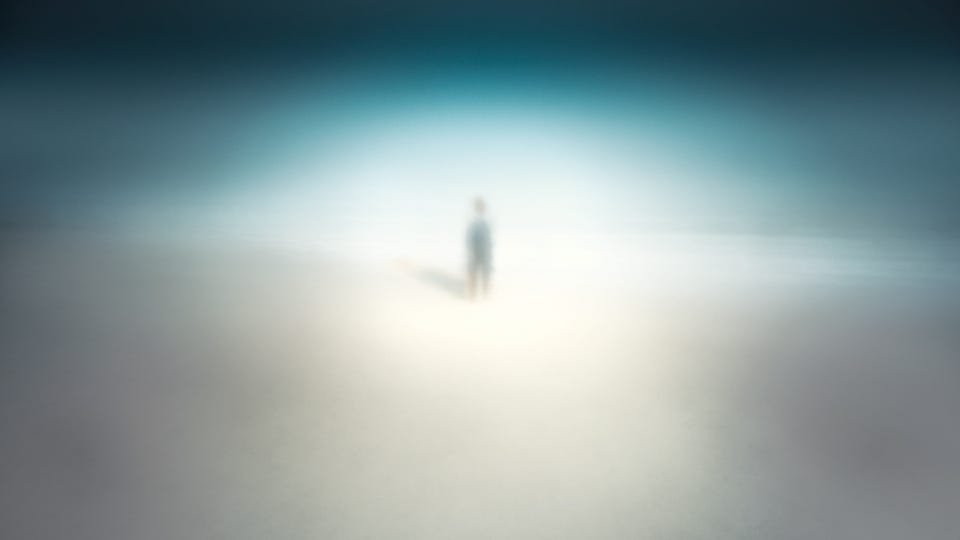 A solitary figure stands small within a soft, glowing haze of white and pale blue, surrounded by open, empty space.