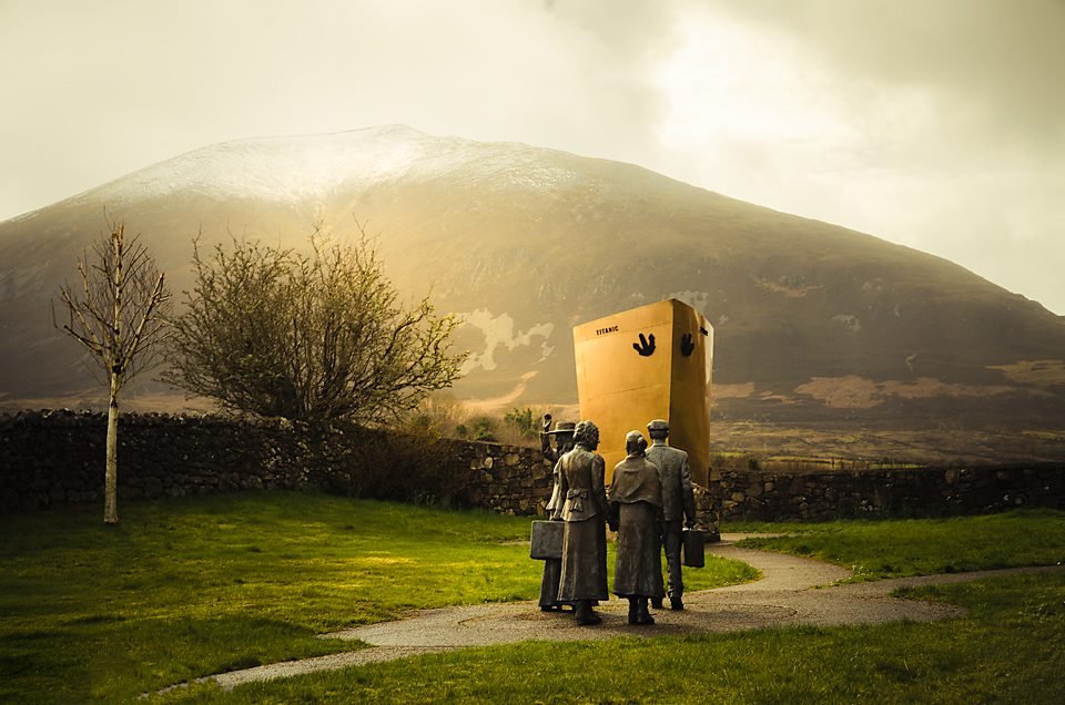 Bronze emigrant statues facing a yellow Titanic memorial sculpture, with a mountain in mist behind, County Mayo.