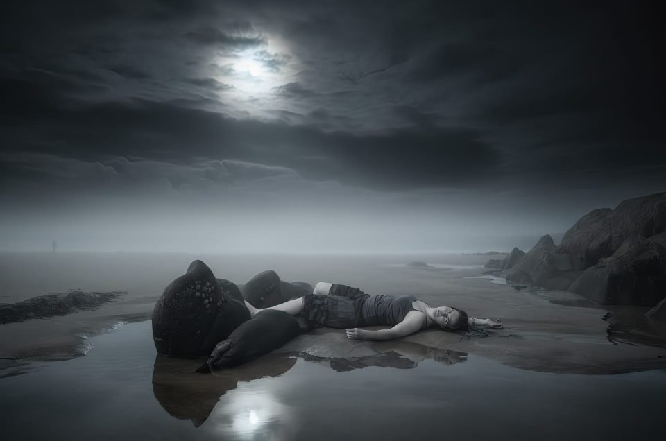A woman lies motionless among dark rocks on wet sand, reflected in shallow water, beneath a stormy sky with the moon breaking through clouds, a distant figure faintly visible on the shoreline.