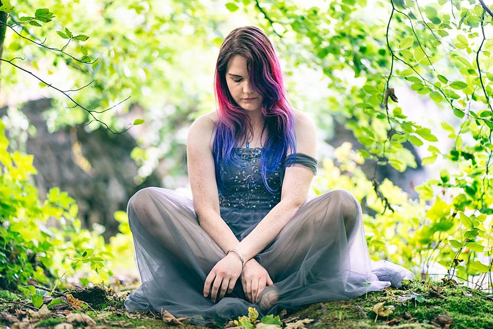 A young woman with pink and blue ombré hair sits cross-legged in a sheer grey dress on mossy ground, surrounded by bright green foliage.