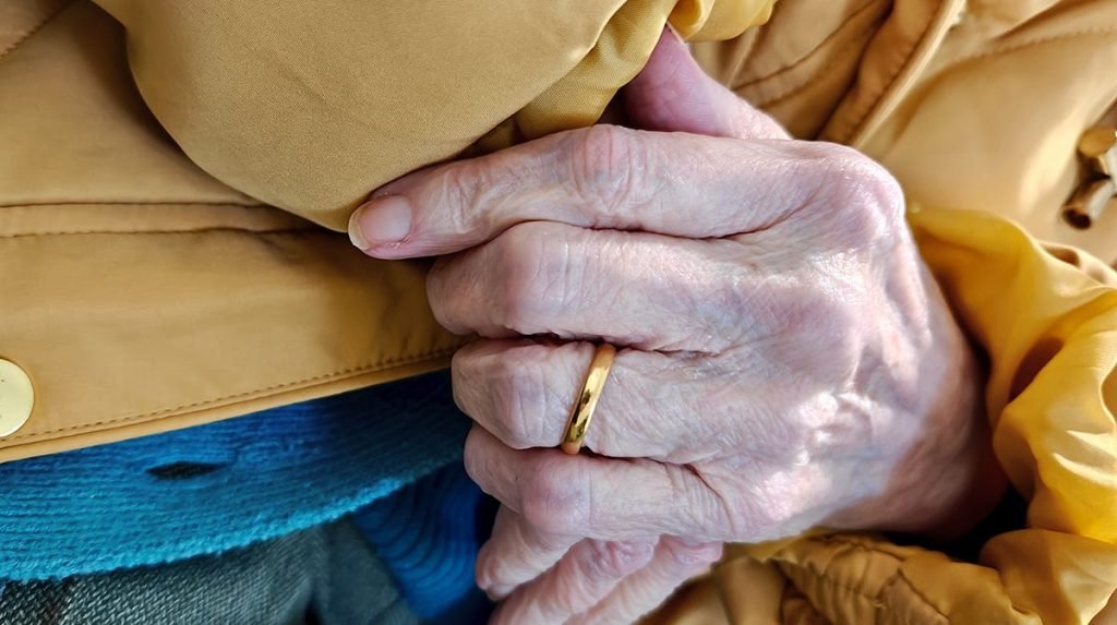 Close-up of clasped, weathered hands with a gold wedding band, resting against a mustard-yellow jacket and blue fabric beneath.